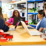 Grupo diverso de estudantes participa de uma aula técnica colaborativa em uma oficina.