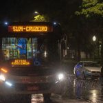 Ônibus de transporte coletivo urbano circula à noite sob chuva em uma avenida.