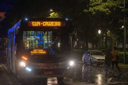 Ônibus de transporte coletivo urbano circula à noite sob chuva em uma avenida.