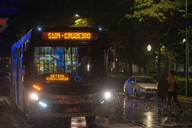 Ônibus de transporte coletivo urbano circula à noite sob chuva em uma avenida.