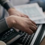 Mãos de uma pessoa conferindo documentos financeiros ao lado de um computador portátil.