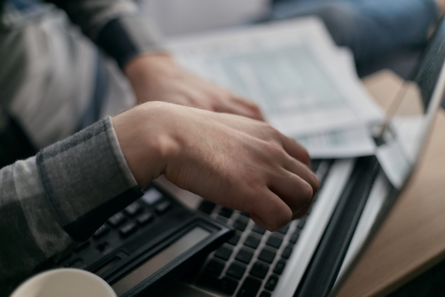 Mãos de uma pessoa conferindo documentos financeiros ao lado de um computador portátil.